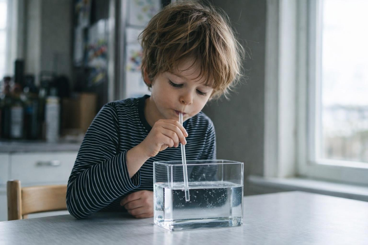 Young boy drinking water from a glass with a straw at a table